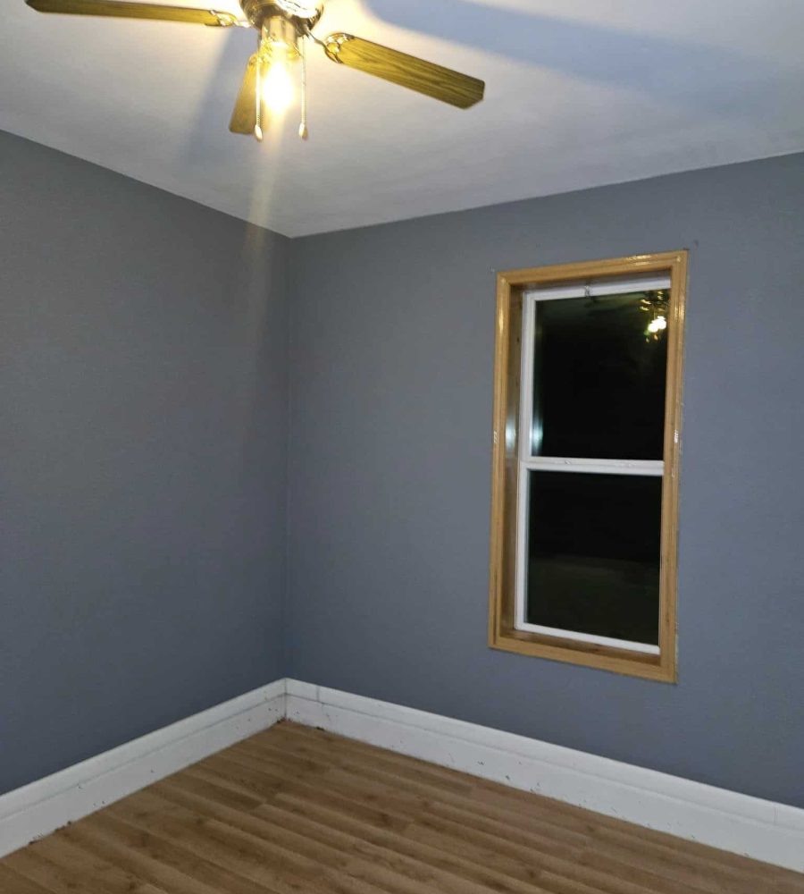 Modern interior room with freshly painted gray walls, a ceiling fan with wooden blades, new wooden window trim, and hardwood flooring, showcasing home improvement and remodeling.