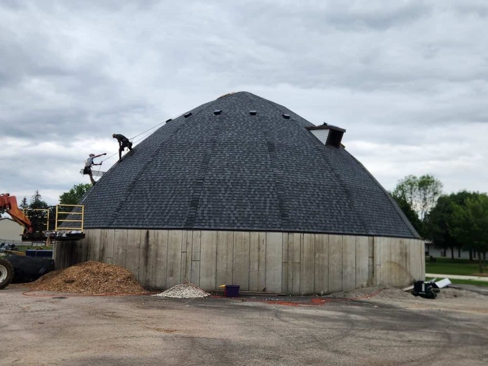 Safe roof replacement with durable shingles on a striking dome-shaped house during daytime. Skilled workers and machinery are used for construction, emphasizing reliable home improvement services.