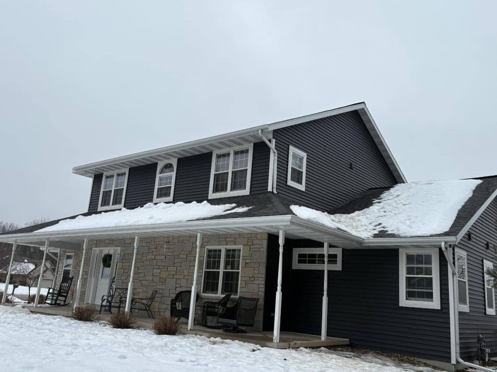 Beautiful two-story house featuring black siding and stone accents, with a snow-covered roof and a spacious porch, showcasing winter residential architecture.